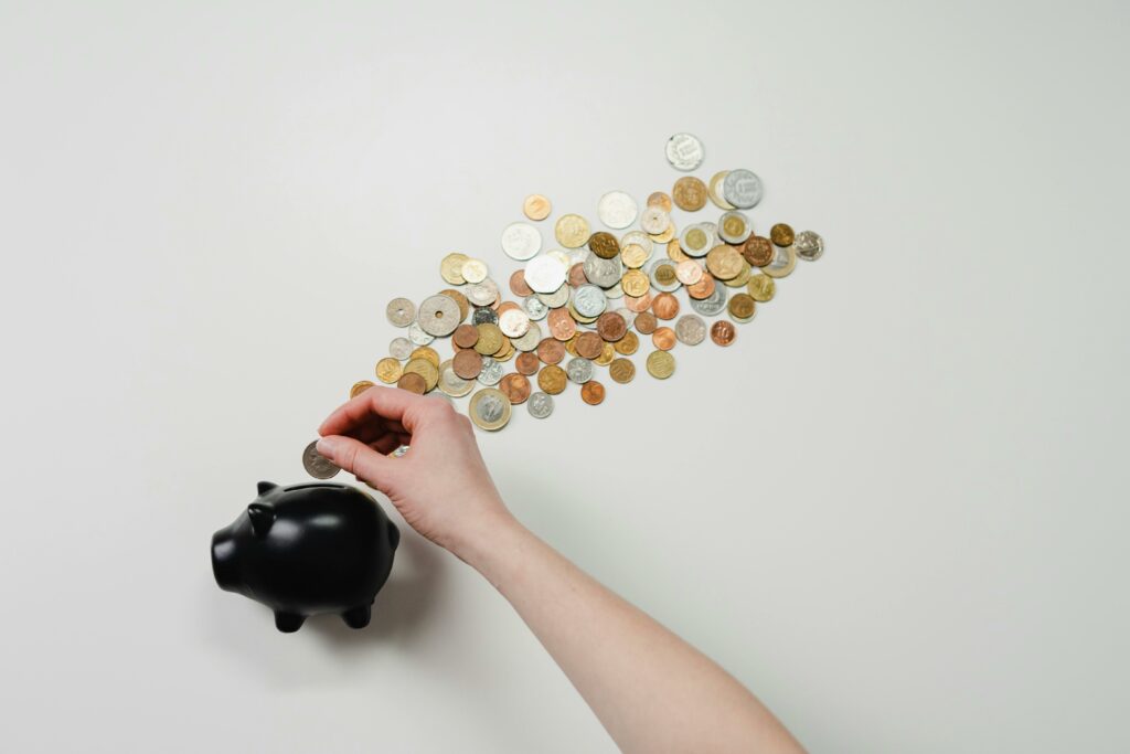 A hand holding a coin over a black piggy bank, with a scattered array of international coins forming an upward trail on a white background.