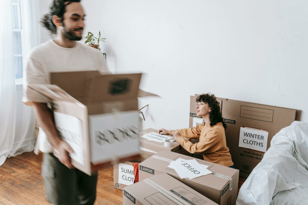A couple in the midst of moving, with cardboard boxes labeled 'Books' and 'Winter Cloths'
