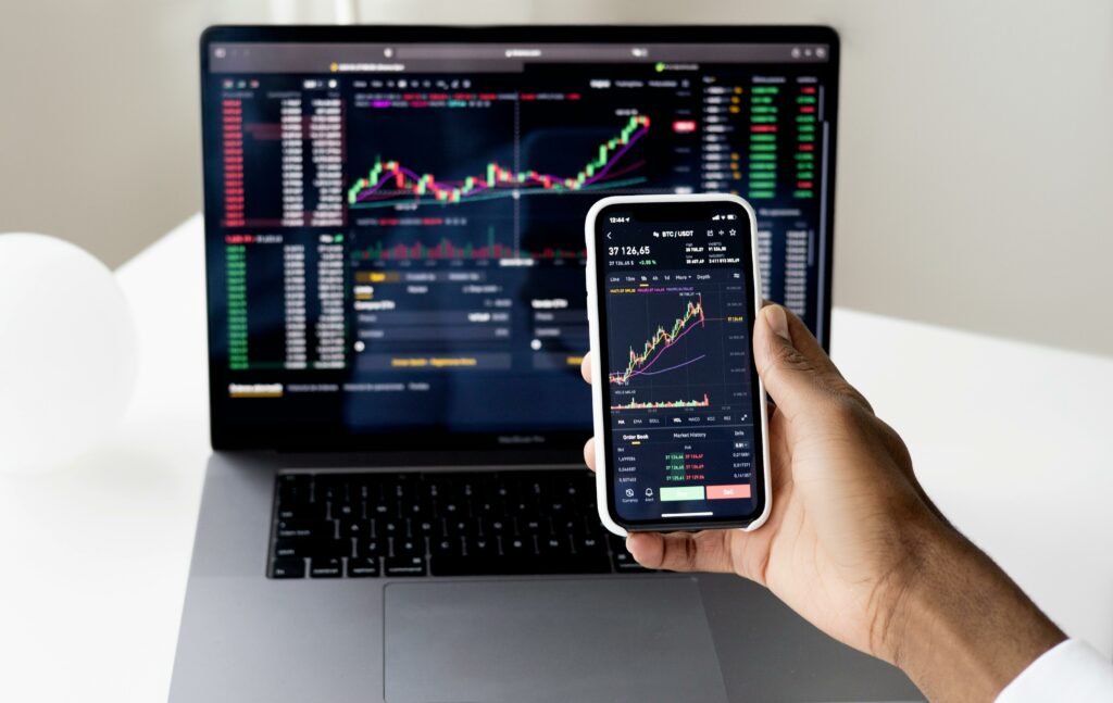 A hand holding a smartphone with stock graphs in front of a laptop with financial data
