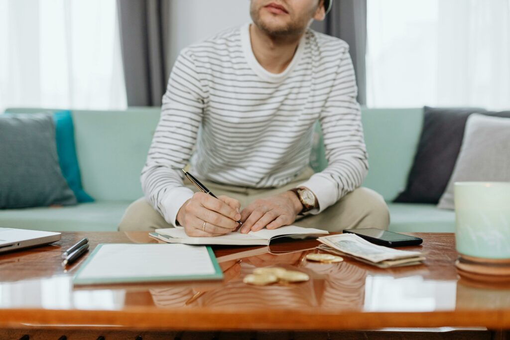 Man sitting at a coffee table focused on writing in a notebook, surrounded by financial planning documents