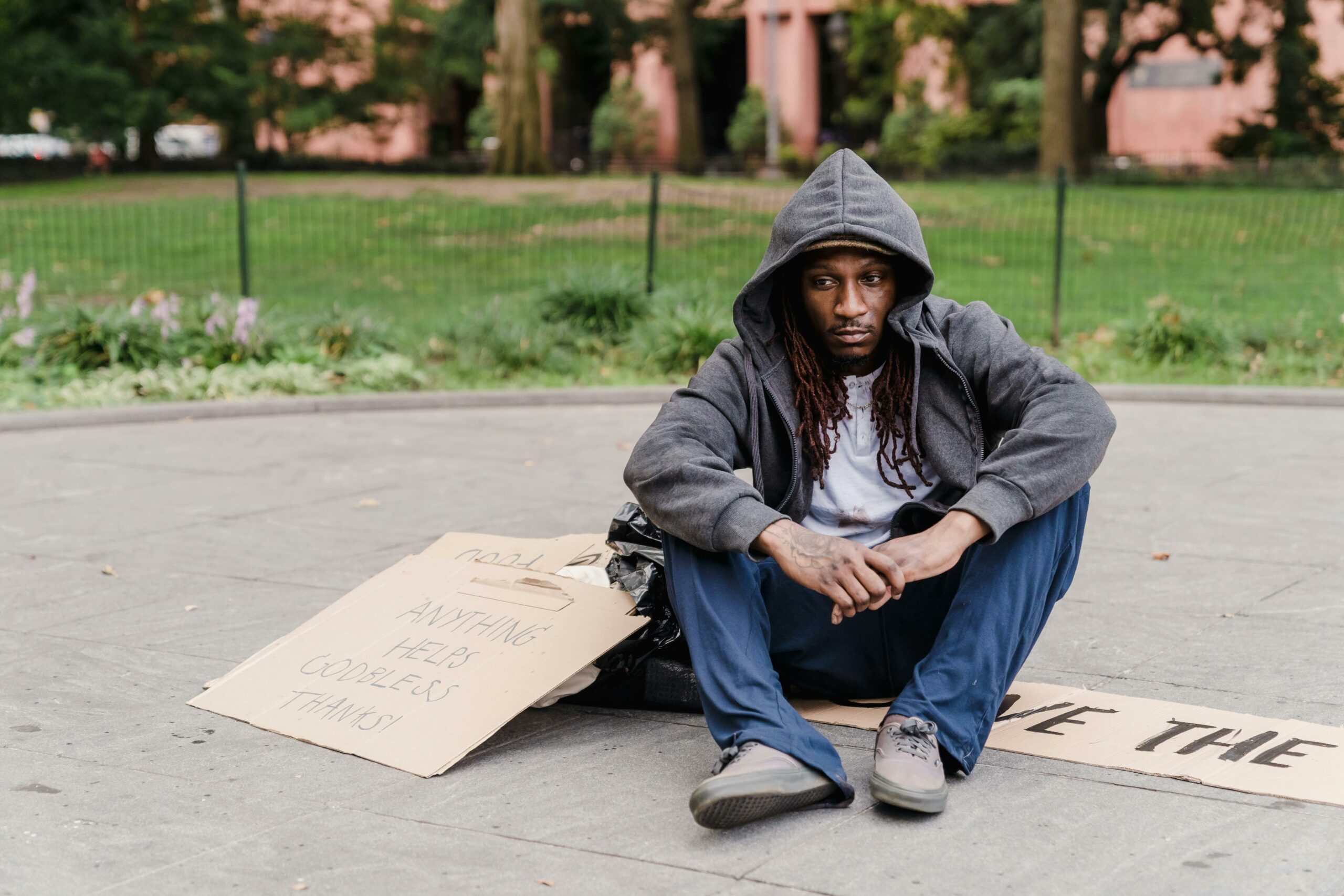 A person sitting on the ground in a park with a sign asking for help, representing the struggle with homelessness.