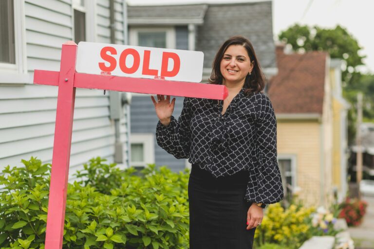 Smiling woman in professional attire standing by a 'SOLD' sign in front of a residential home.