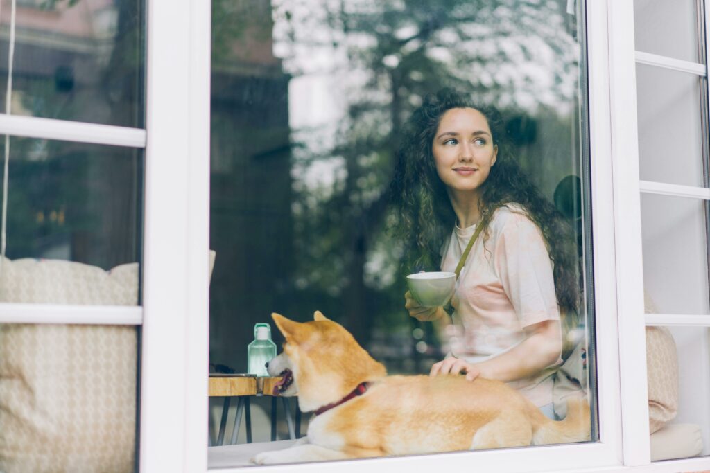 Young woman with curly hair and a Shiba Inu dog by the window, reflecting on the benefits of guarantor home loans.