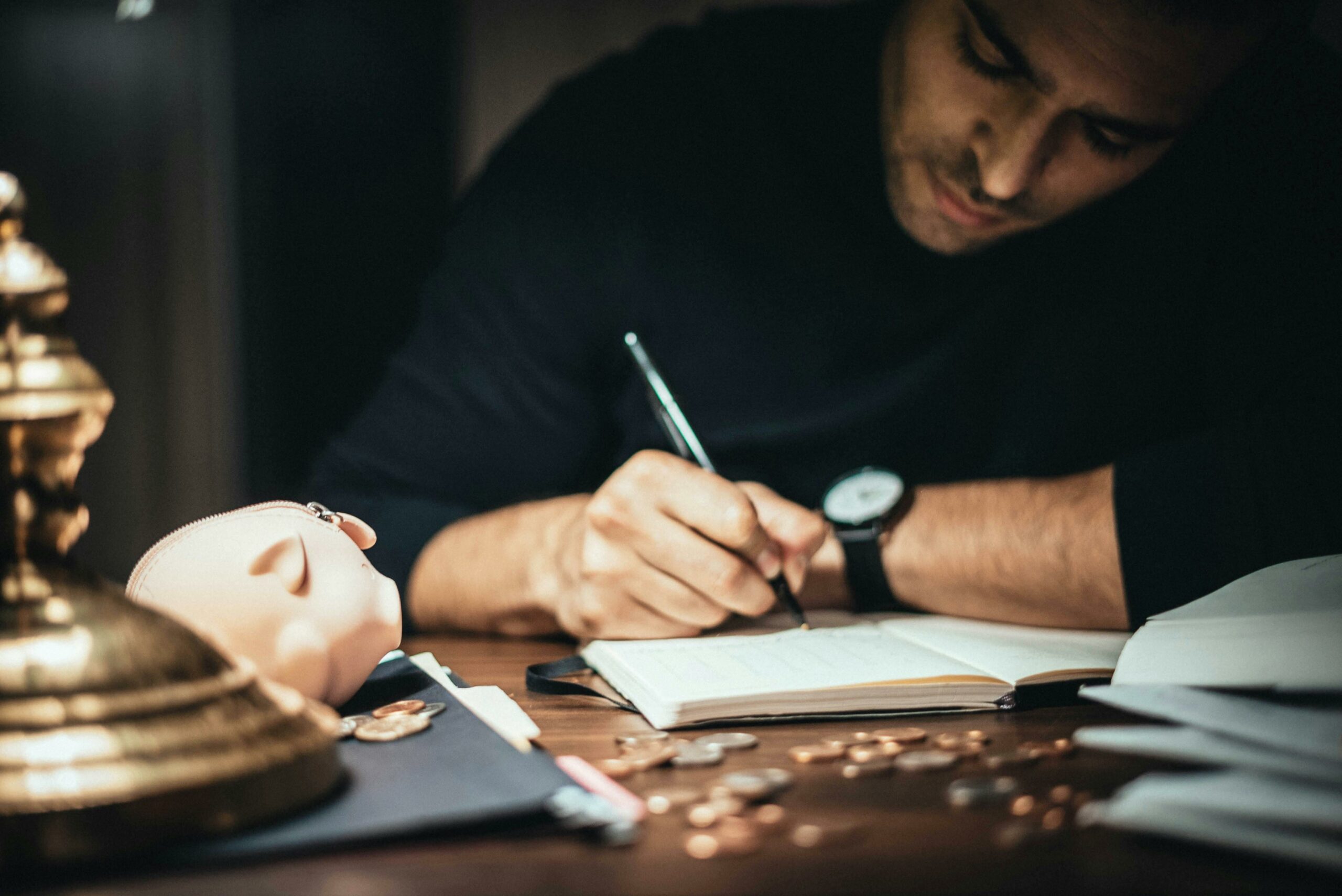 A man writing in a notebook at a desk, with a piggy bank, coins, and papers spread around him, focusing intently on his work.