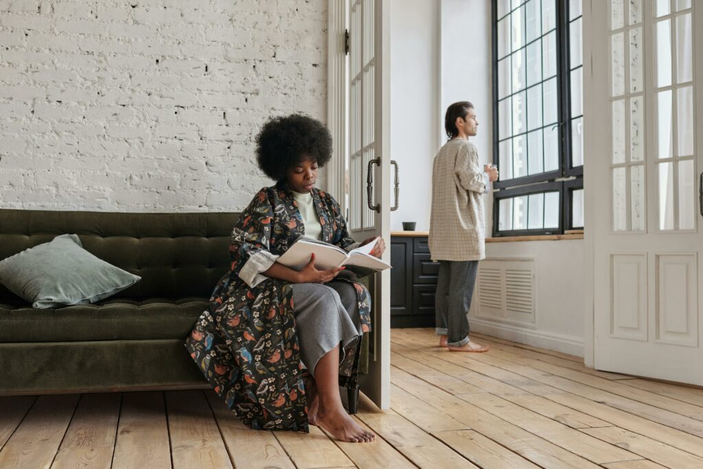 Woman reading a book on a couch while a man looks out the window in a cozy, modern apartment.