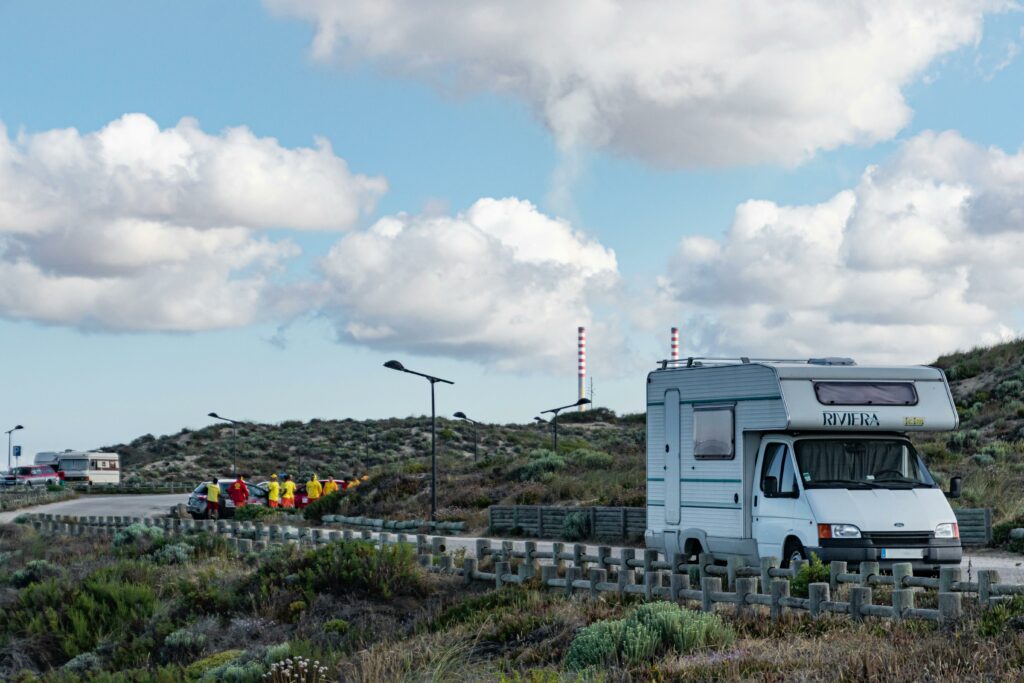A caravan parked on a scenic roadside with a group of people in bright clothing gathered nearby, under a blue sky with fluffy clouds.