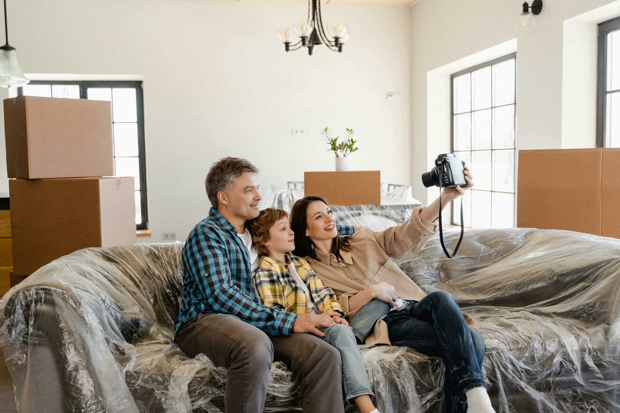 A family of three sitting on a couch covered in plastic wrap, taking a selfie in their new home with moving boxes in the background.