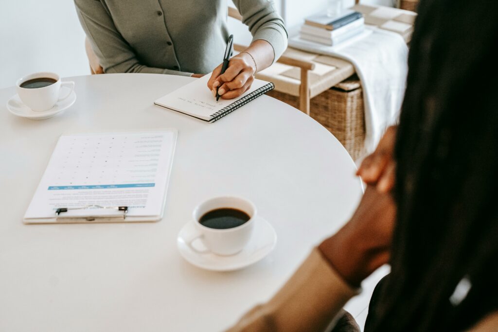 Two people sitting at a table with coffee cups, a notepad, and a clipboard, engaged in a discussion.