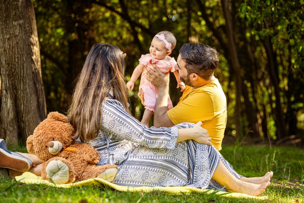 A young family enjoying a picnic in the park, with the father holding up a smiling baby, the mother sitting beside them, and a large teddy bear resting on a blanket.