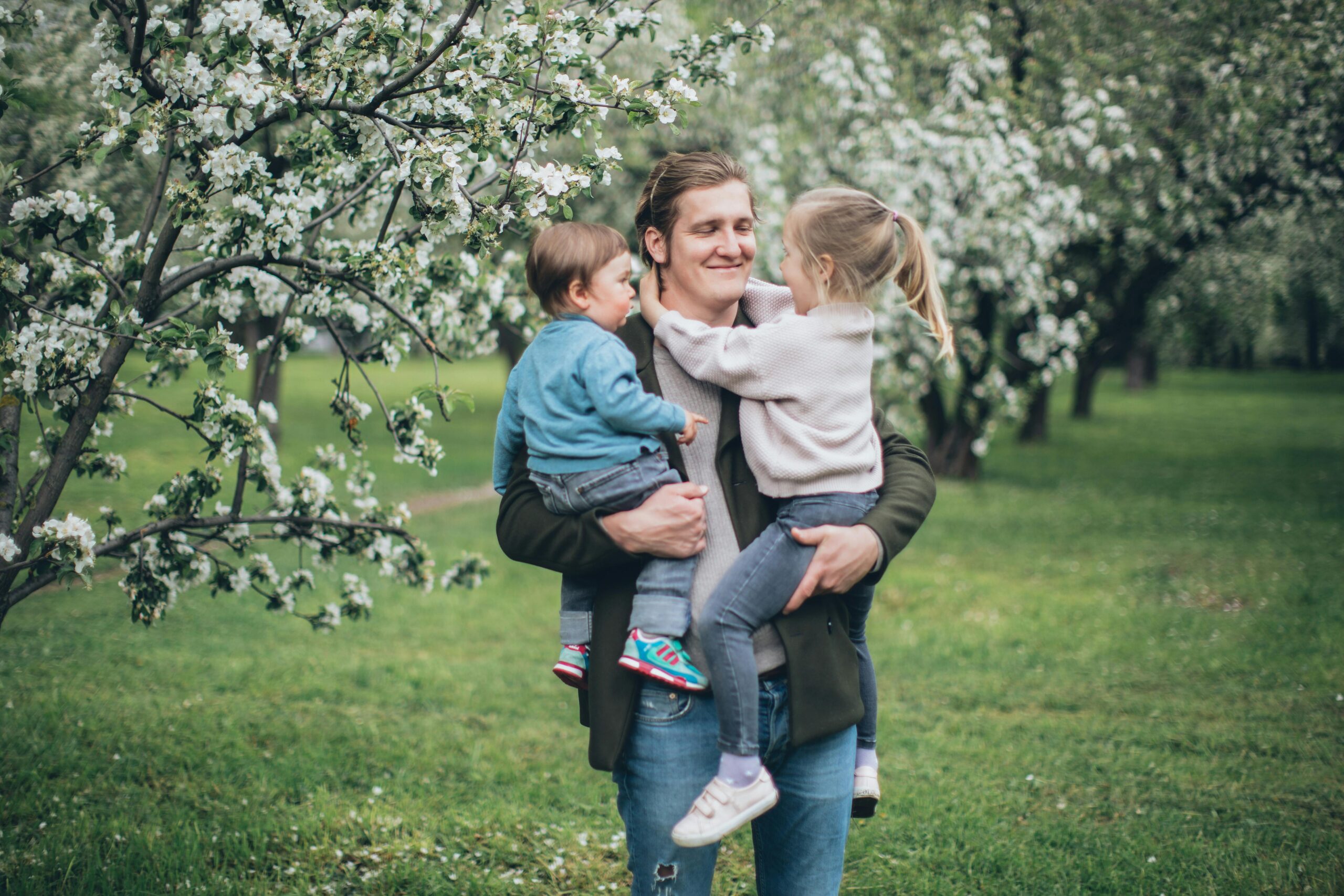 A man holding two young children, one in each arm, standing in an orchard with blooming trees. The children are smiling and interacting with the man, creating a warm and affectionate scene in a natural setting.