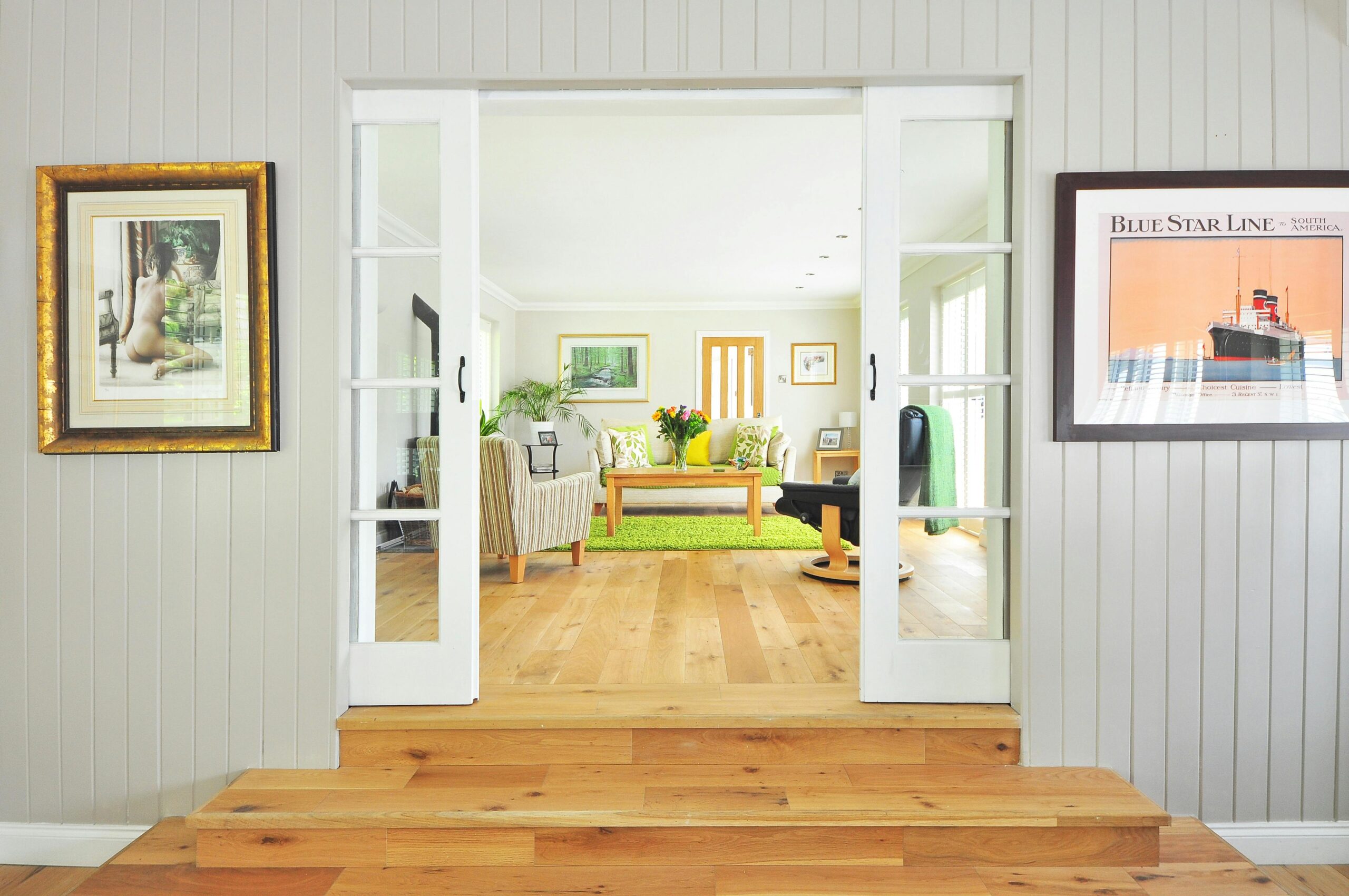 Interior of a bright, modern living room viewed through open sliding glass doors, featuring light-colored furniture, a green rug, and framed artwork on the walls