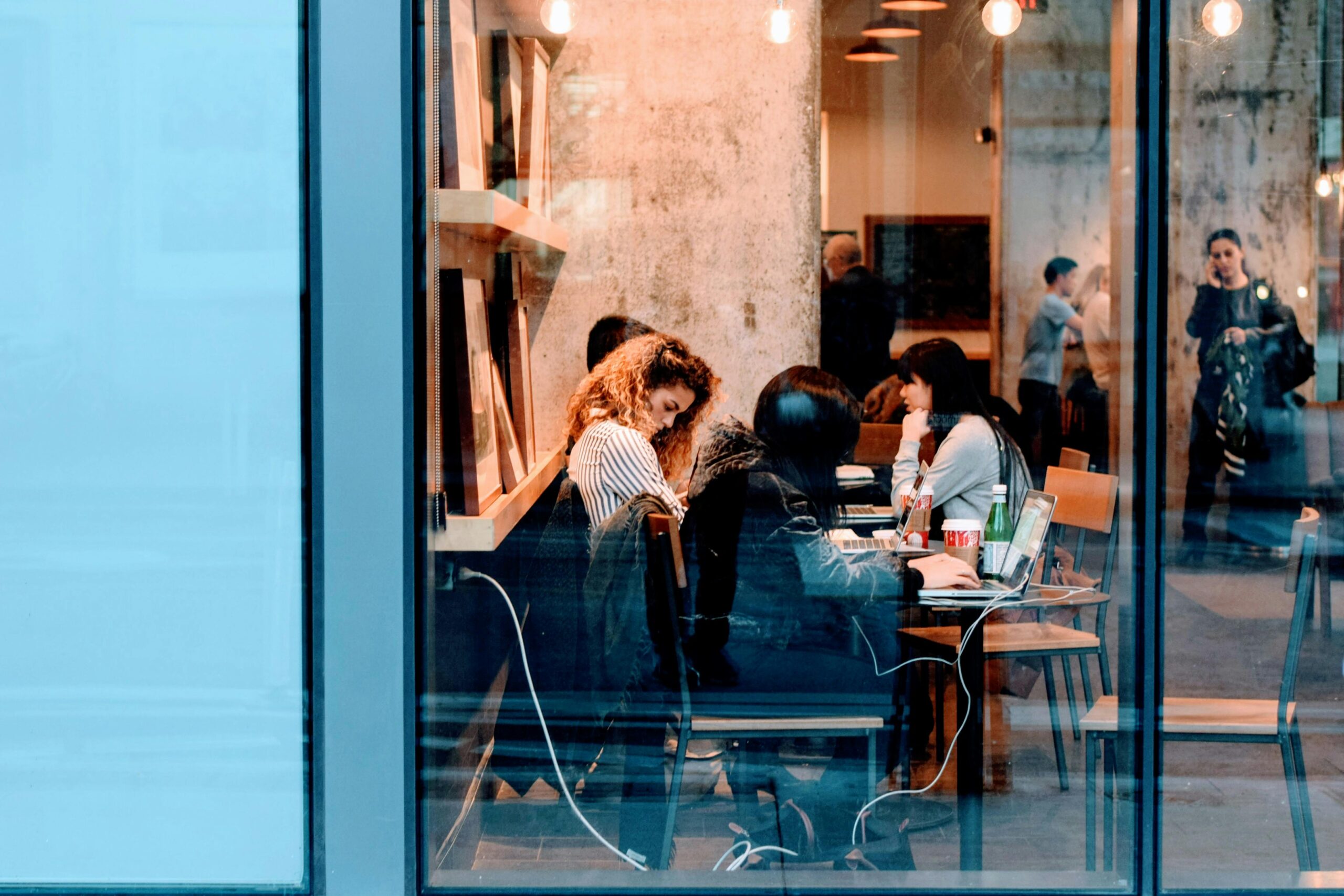 A group of people working on laptops in a modern cafe, viewed through a large glass window.