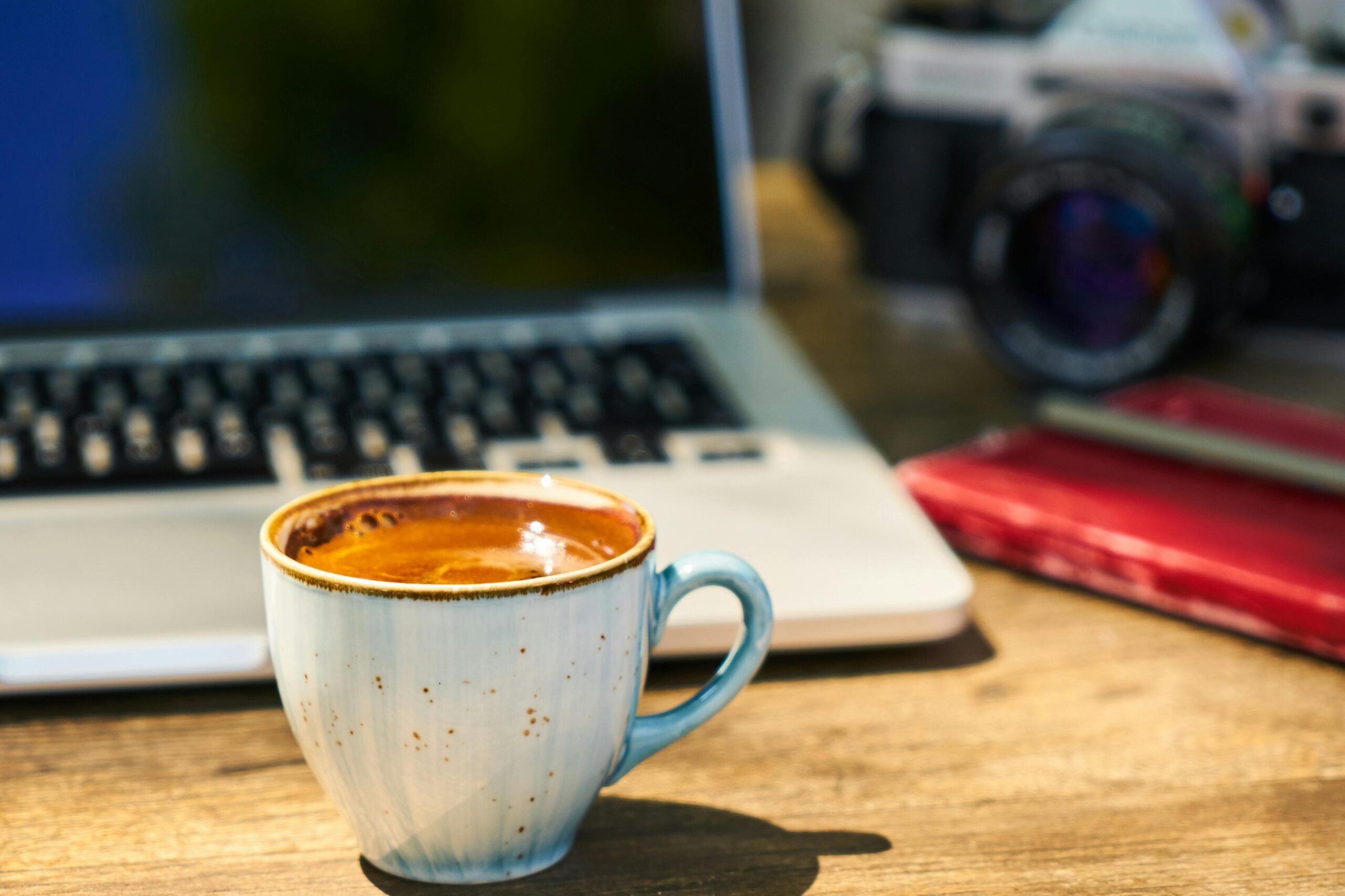 Close-up of a ceramic coffee cup filled with espresso on a wooden table beside a laptop and camera.