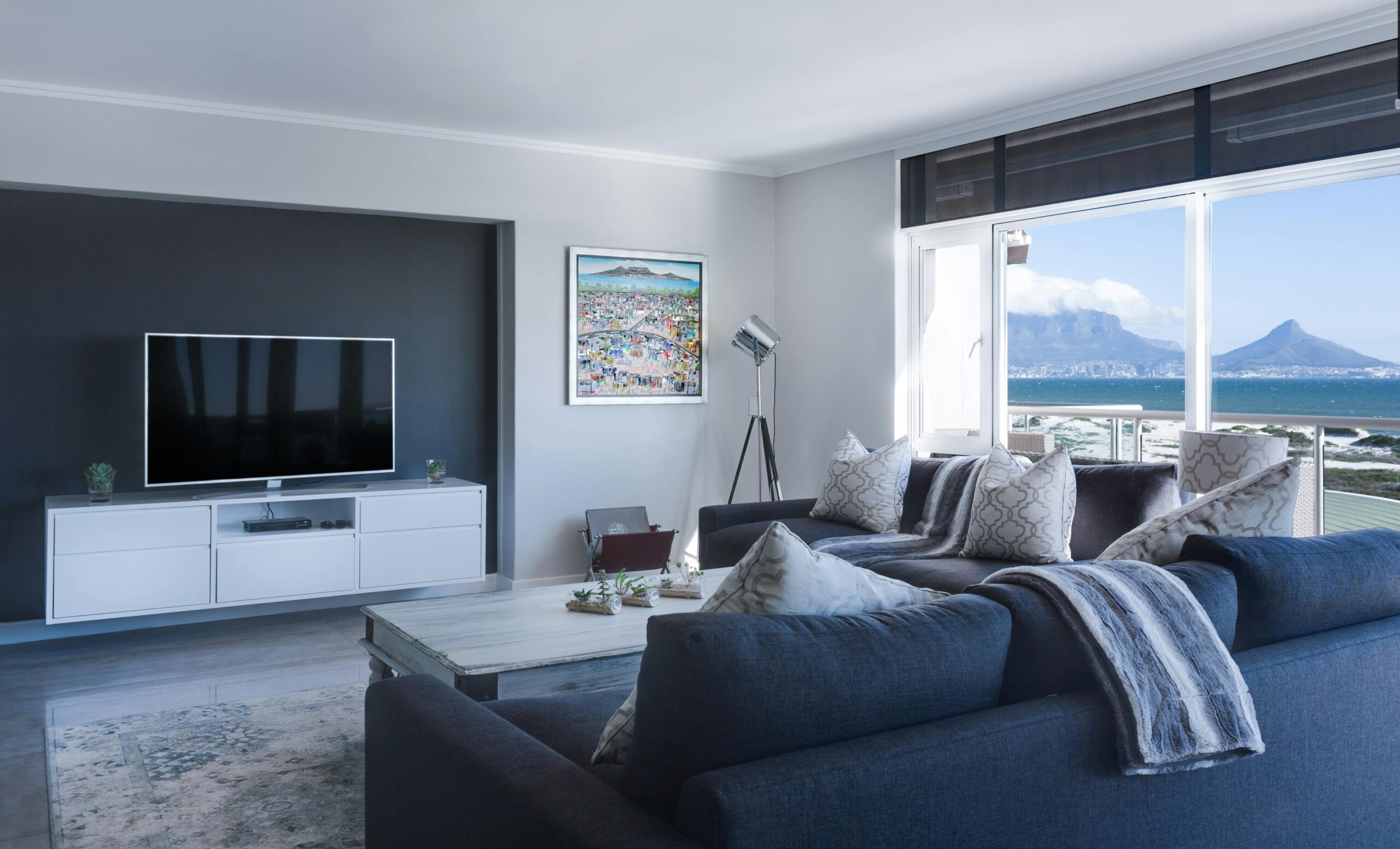 Modern living room with large windows offering a scenic view of mountains and the ocean. The room features a grey sofa, a coffee table, and a wall-mounted TV.