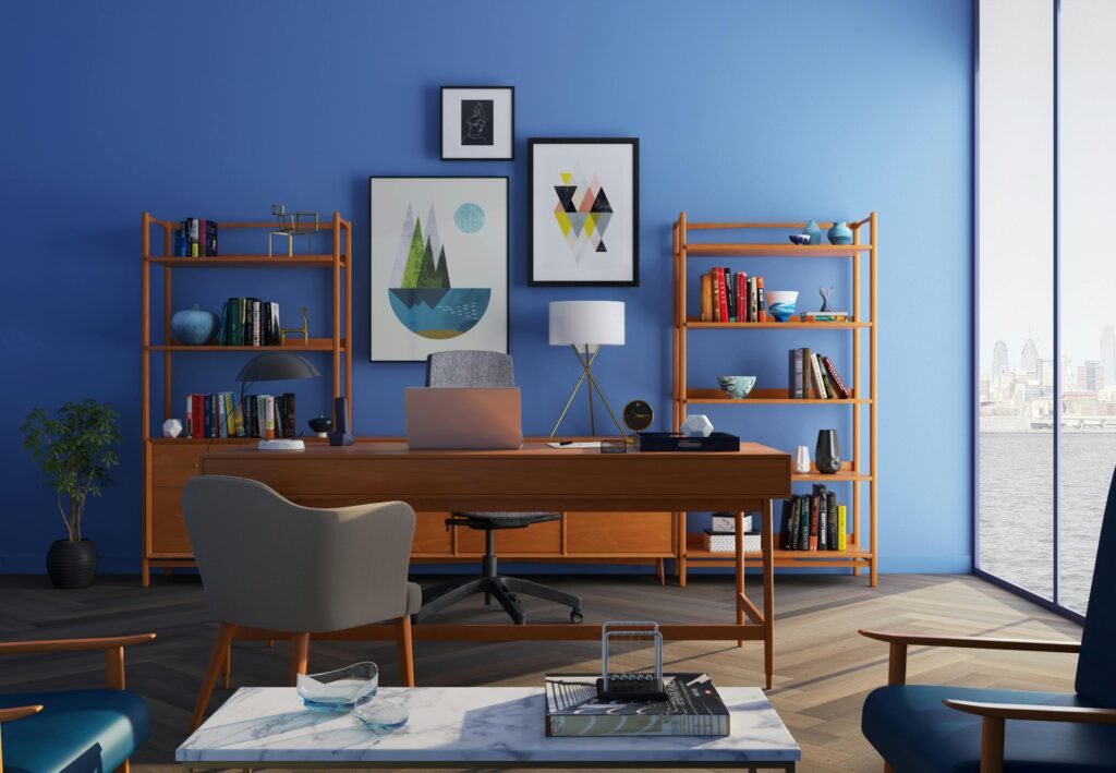 Modern home office setup with a wooden desk, bookshelf, and framed artwork on a blue wall, overlooking a cityscape through large windows.