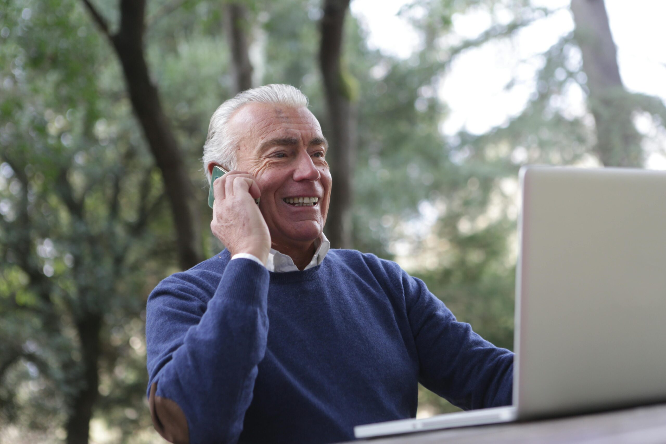 Smiling elderly man in a blue sweater holding a smartphone to his ear while using a laptop outdoors.