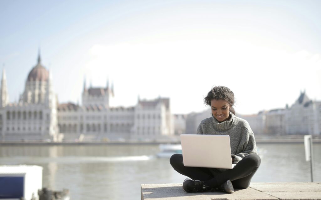Young woman sitting outdoors with a laptop on her lap, smiling while working, with a blurred view of a large historic building and river in the background.