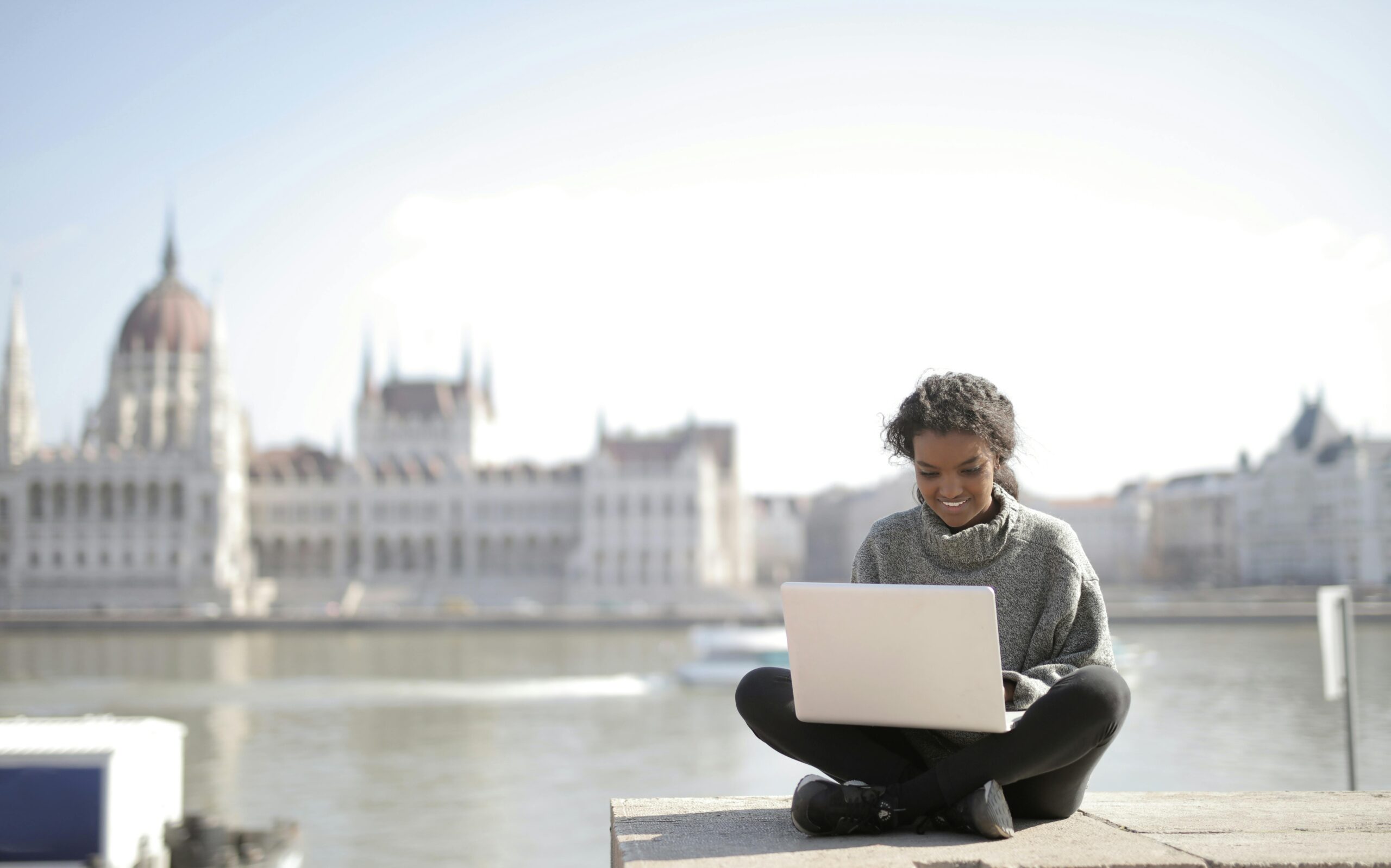 Young woman sitting outdoors with a laptop on her lap, smiling while working, with a blurred view of a large historic building and river in the background.